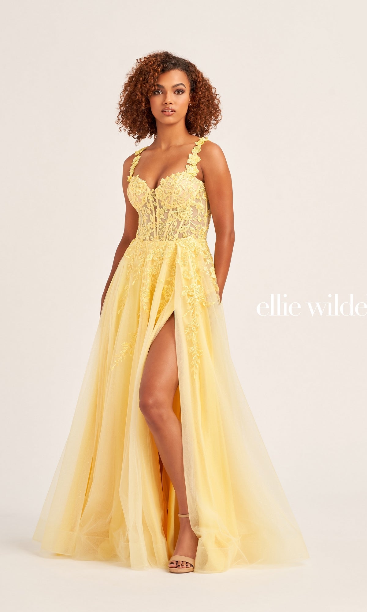 A woman wearing the Ellie Wilde Off-Shoulder Long Prom Dress EW35101— a yellow glitter tulle ball gown with a fitted bodice, floral accents, and a high slit—stands against a neutral background.